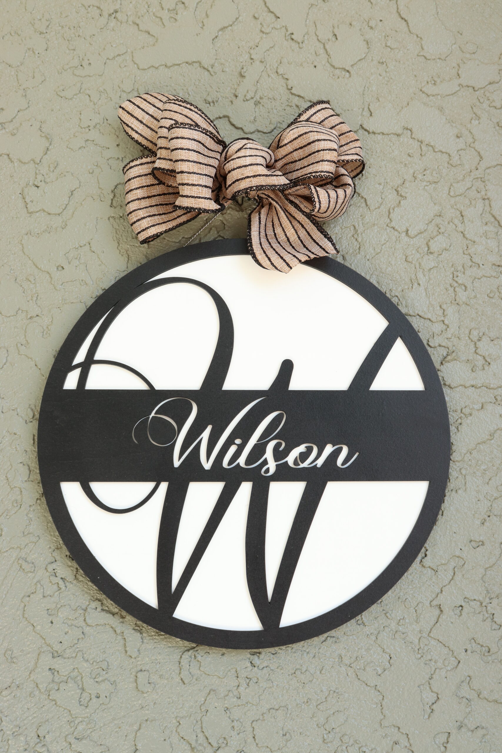 Wilson Initial Sign This round sign is laser cut with a large, raised "W" initial in black with a border and central cross banner in black. The white background allows that initial and the laser cut "Wilson" recess in the cross piece to pop against the black. It is topped with a black striped burlap bow as an accent.