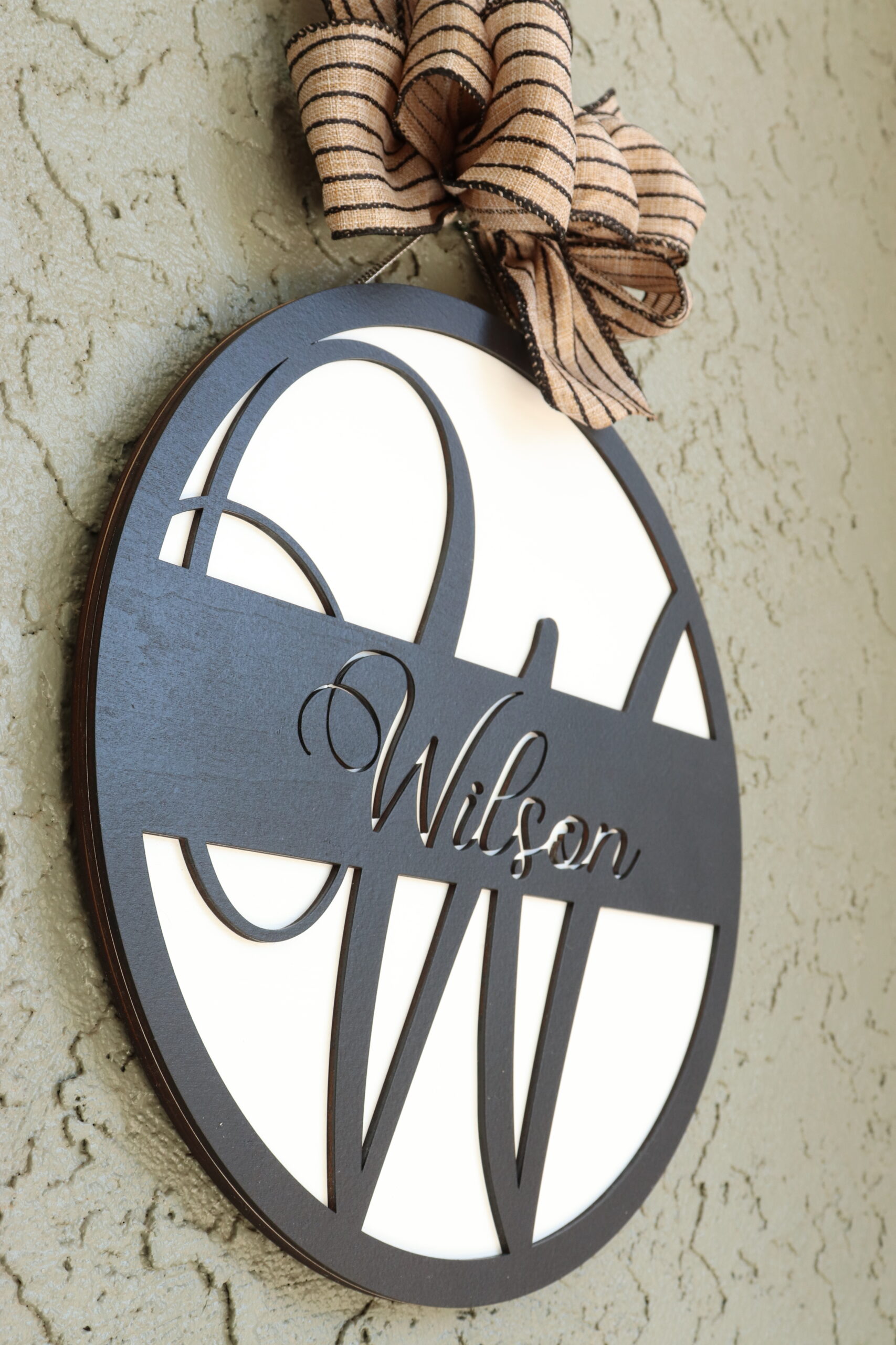 Wilson Initial Sign Side View This round sign is laser cut with a large, raised "W" initial in black with a border and central cross banner in black. The white background allows that initial and the laser cut "Wilson" recess in the cross piece to pop against the black. It is topped with a black striped burlap bow as an accent.